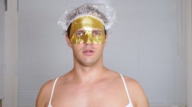 Yüz paketi olan garip adam. Genç bir adam bir ev bir ayna önünde yüz için kozmetik işlemler yapar. 4k, altın maske