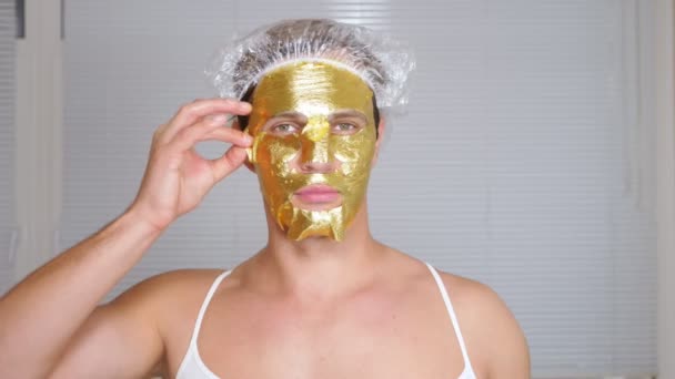 Strano uomo con la faccia piena. Un giovane fa procedure cosmetiche per il volto di una casa di fronte a uno specchio. 4k, maschera dorata — Video Stock