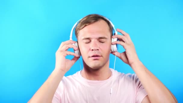 Jeune homme sur un fond bleu coloré. portrait. émotions et gestes. 4k, gros plan. Au ralenti. gars écoutant de la musique dans des écouteurs blancs — Video