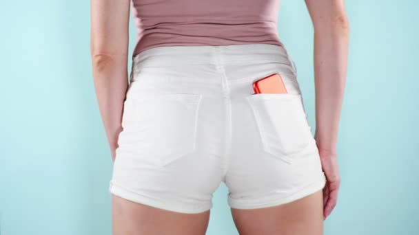 Mulher elegante com um smartphone em seus shorts de bolso. Moda de verão. Férias. 4k, câmera lenta, fundo de cor . — Vídeo de Stock