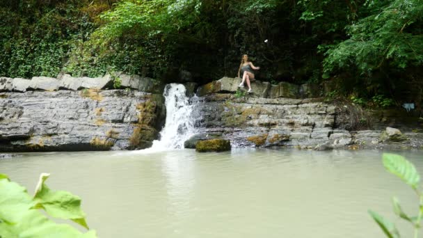 Blogueira está liderando um relatório sentado em uma rocha perto da cachoeira. A natureza pitoresca de uma bela cachoeira e uma esmeralda de um lago de água doce no ambiente florestal de selvas selvagens. 4k — Vídeo de Stock