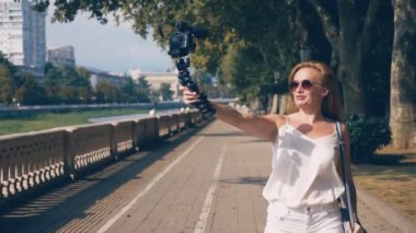 güzel mutlu kadın blogger bir kamera ile ona röportaj resort şehir set strolling yol açar. rüzgar saçlarını ağaçların dalları ile güneş ışığı glintes geliştirir