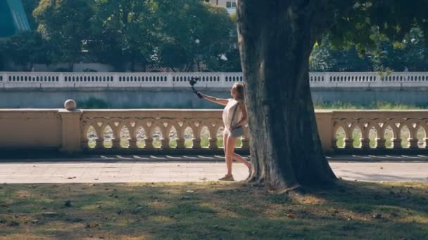 Güzel mutlu kadın blogger bir kamera ile ona röportaj resort şehir set strolling yol açar. rüzgar saçlarını ağaçların dalları ile güneş ışığı glintes geliştirir — Stok video