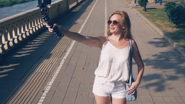 Hermosa bloguera feliz mujer con una cámara, conduce su reportaje paseando por el terraplén de la ciudad turística. el viento desarrolla su cabello, la luz del sol brilla a través de las ramas de los árboles — Vídeos de Stock