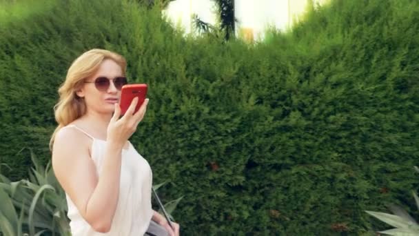 Gelukkig jonge vrouw praten over de telefoon, wandelen langs de waterkant op een warme zonnige zomerdag. 4k, slow-motion — Stockvideo
