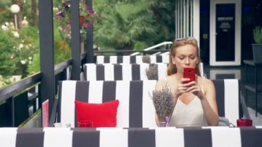 Ayrılık ile mutsuz olmak telefon bakarak kafede masada oturan bir genç bayan. 4k, ağır çekim, şık kafe iç. Siyah ve beyaz çizgili ve kırmızı vurgu