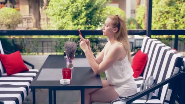 Mujer joven sentada en la mesa en la cafetería mirando el teléfono siendo infeliz con la ruptura. 4k, cámara lenta, elegante interior de la cafetería. Rayas blancas y negras y acento rojo — Vídeos de Stock