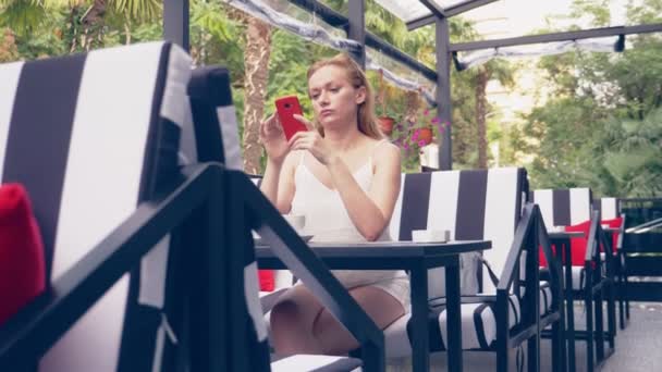 Jovem mulher sentada à mesa no café olhando para o telefone sendo infeliz com a separação. 4k, câmera lenta, interior elegante café. Listras preto e branco e sotaque vermelho — Vídeo de Stock