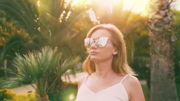 Hermosa mujer rubia con estilo en gafas de sol, caminando a lo largo de un camino de palmeras. La palma se refleja en los vasos. 4K cámara lenta . — Vídeo de stock