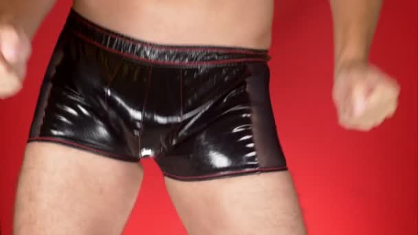 Sexy modelo masculino en ropa interior sobre un fondo rojo. Un hombre con pantalones de látex negro mueve sus caderas. 4k, cámara lenta. primer plano — Vídeo de stock
