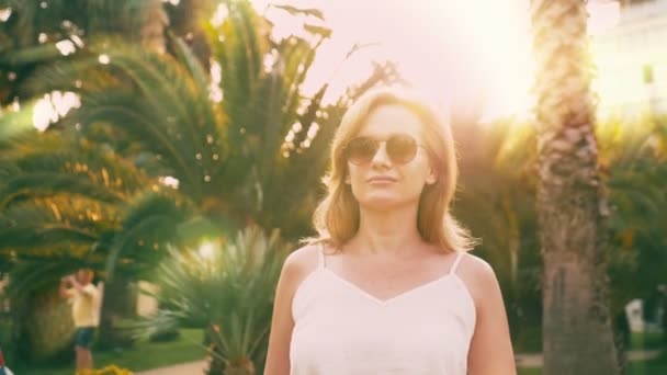 Mulher loira elegante bonita em óculos de sol, andando ao longo de um caminho palmeira. A palma da mão é refletida nos óculos. Movimento lento 4K . — Vídeo de Stock
