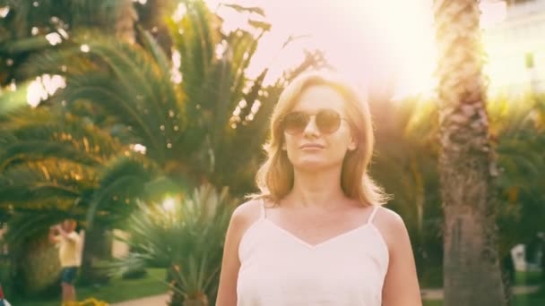 Mooie stijlvolle blonde vrouw in zonnebril, wandelen langs een palm tree pad. De palm wordt weerspiegeld in de glazen. 4 k slow-motion. — Stockvideo