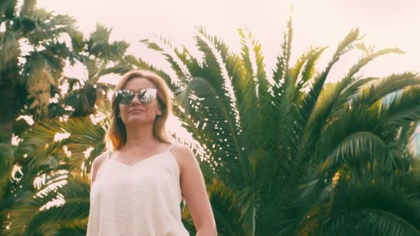 Hermosa mujer rubia con estilo en gafas de sol, caminando a lo largo de un camino de palmeras. La palma se refleja en los vasos. 4K cámara lenta . — Vídeo de stock