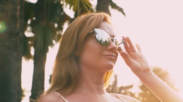 Bir palmiye ağacı yol boyunca yürüyüş güzel şık sarışın kadın güneş gözlüğü. Palmiye gözlükte yansıtılır. 4 k yavaş hareket. — Stok video