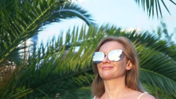 Belle femme blonde élégante en lunettes de soleil, marchant le long d'un sentier de palmiers. La paume se reflète dans les lunettes. 4K au ralenti . — Video
