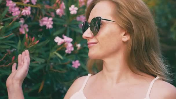 Hermosa mujer rubia con estilo en gafas de sol caminando en el parque con arbustos florecientes. flores de adelfa se reflejan en los vasos. 4K cámara lenta . — Vídeo de stock