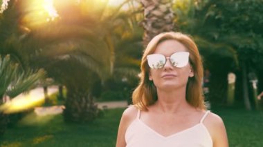 Bir palmiye ağacı yol boyunca yürüyüş güzel şık sarışın kadın güneş gözlüğü. Palmiye gözlükte yansıtılır. 4 k yavaş hareket.