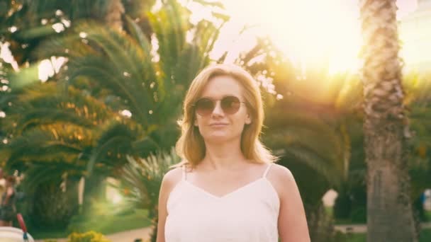 Hermosa mujer rubia con estilo en gafas de sol, caminando a lo largo de un camino de palmeras. La palma se refleja en los vasos. 4K cámara lenta . — Vídeo de stock