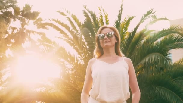 Bella donna bionda elegante in occhiali da sole, passeggiando lungo un sentiero di palme. Il palmo si riflette nei bicchieri. 4K rallentatore . — Video Stock