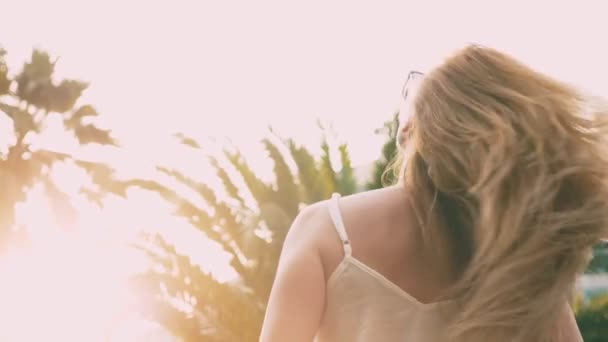 Belle femme blonde élégante en lunettes de soleil, marchant le long d'un sentier de palmiers. La paume se reflète dans les lunettes. 4K au ralenti . — Video