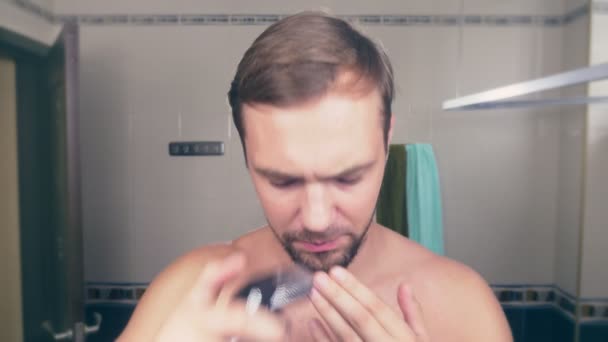 A handsome young man shaves his beard with an electric shaver, standing in the bathroom in front of the mirror. 4k, slow motion — Stock Video