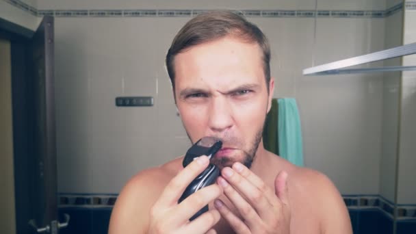 Un beau jeune homme se rase la barbe avec un rasoir électrique, debout dans la salle de bain devant le miroir. 4k, au ralenti — Video