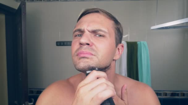 A handsome young man shaves his beard with an electric shaver, standing in the bathroom in front of the mirror. 4k, — Stock Video