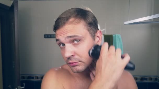 Um jovem bonito rapa a barba com um barbeador elétrico, de pé no banheiro em frente ao espelho. 4k , — Vídeo de Stock