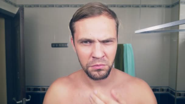 Красивий молодий чоловік голить бороду електричною бритвою, стоячи у ванній перед дзеркалом. 4k, повільний рух — стокове відео