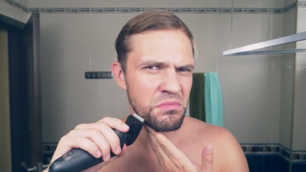 Un beau jeune homme se rase la barbe avec un rasoir électrique, debout dans la salle de bain devant le miroir. 4k, au ralenti — Video