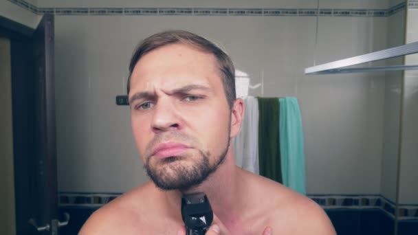 Un beau jeune homme se rase la barbe avec un rasoir électrique, debout dans la salle de bain devant le miroir. 4k, au ralenti — Video