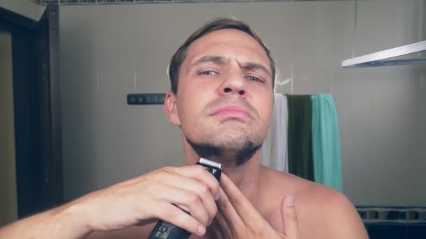 Um cara bonito engraçado barba sua barba com um barbeador elétrico no banheiro e canta canções na frente de um espelho. 4k . — Vídeo de Stock