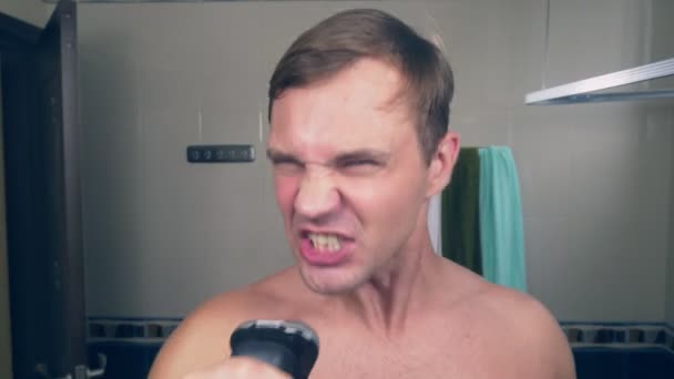 En rolig stilig kille rakar skägget med en rakapparat i badrummet och sjunger låtar framför en spegel. 4k. — Stockvideo