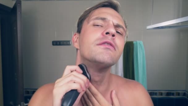 Un beau mec drôle se rase la barbe avec un rasoir électrique dans la salle de bain et chante des chansons devant un miroir. 4k . — Video