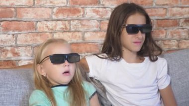 Kapatın. iki küçük kız bardaklarda 3d, film izlerken çok duygu ile oturma odasında kanepede oturuyor. 4k, ağır çekim.