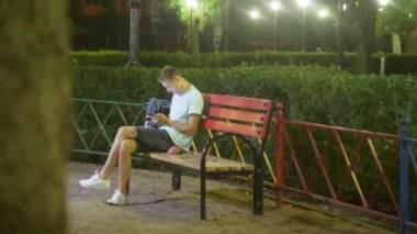 Bir akıllı telefon ile bir bankta oturan çekici üzgün adam. sıcak yaz gecesi geç. parkta. 4k, steadicam vurdu