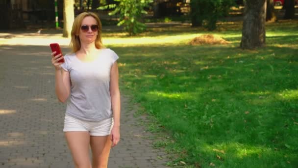 Kvinna kommer till parken med sin telefon på sommardag under solen ljus. 4 k, slow motion, standicam skott — Stockvideo