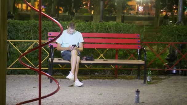 Привабливий сумний чоловік сидить на лавці зі смартфоном. пізно в теплу літню ніч. в парку. 4k, стайка постріл — стокове відео