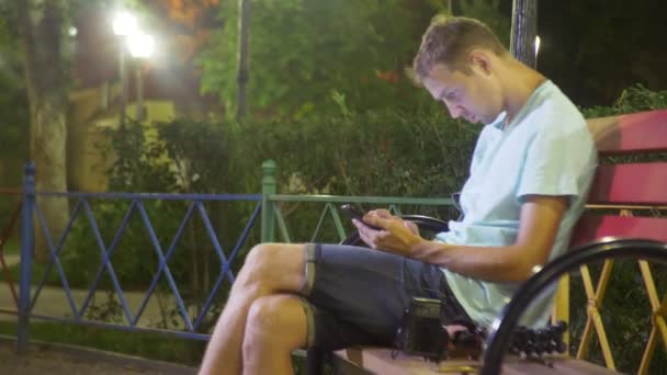 Atractivo hombre triste sentado en un banco con un teléfono inteligente. tarde en una cálida noche de verano. en el parque. 4k, disparo steadicam — Vídeo de stock