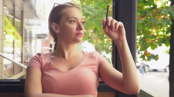 Une jeune femme fume de fines cigarettes alors qu'elle est assise sur une véranda dans un café, un matin d'été ensoleillé. 4k, au ralenti — Video