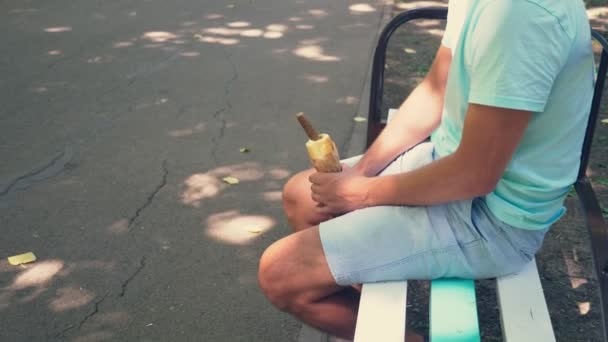 Un giovane ragazzo solitario che tiene in braccio un hotdog, nell'addome inferiore, seduto su una panchina nel parco, e apre la confezione dell'hotdog, imitando un'erezione. 4k, rallentatore — Video Stock