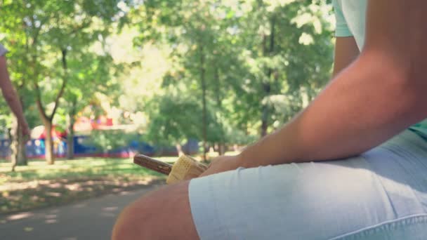 Un jeune homme seul tenant un hot-dog, dans le bas-ventre, assis sur un banc dans le parc, et ouvrant l'emballage du hot-dog, imitant une érection. 4k, au ralenti — Video