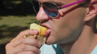 güneş gözlüğü, bir şehir parkta bir bankta oturan zevkle sosisli sandviç yeme adam. 4k, ağır çekim
