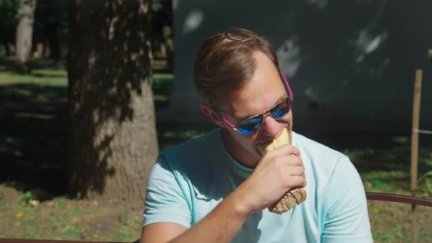 Mann mit Sonnenbrille, der genüsslich Hot Dogs isst und im Stadtpark auf einer Bank sitzt. 4k, Zeitlupe — Stockvideo