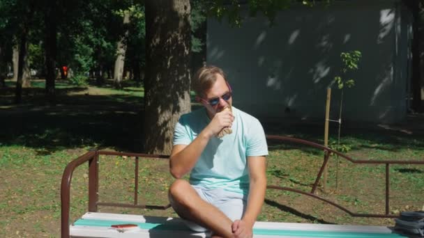 Mann mit Sonnenbrille, der genüsslich Hot Dogs isst und im Stadtpark auf einer Bank sitzt. 4k, Zeitlupe — Stockvideo