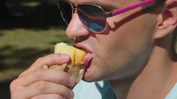 Хлопець в сонцезахисних окулярах, з задоволенням їсть хот-доги, сидячи в міському парку на лавці. 4k, повільний рух — стокове відео