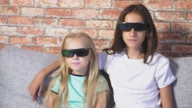 Kapatın. iki küçük kız bardaklarda 3d, film izlerken çok duygu ile oturma odasında kanepede oturuyor. 4k, ağır çekim.
