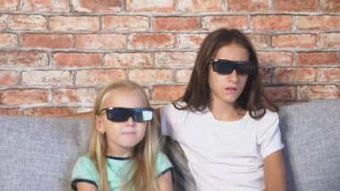Kapatın. iki küçük kız bardaklarda 3d, film izlerken çok duygu ile oturma odasında kanepede oturuyor. 4k, ağır çekim.