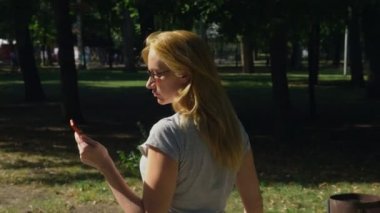 Yaz günü güneş ışığı altında onun telefon ile park olacak kadın. 4 k, yavaş hareket, standicam atış