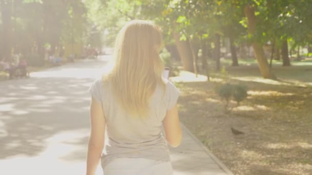 Kvinna kommer till parken med sin telefon på sommardag under solen ljus. 4 k, slow motion, standicam skott — Stockvideo